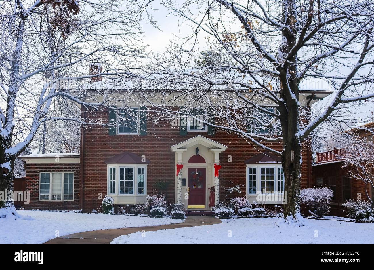 Belle maison en brique avec baies vitrées avec arbre de Noël montrant à travers et décoré des piliers et traîneau sur le porche dans la neige encadrée par des arbres d'hiver Banque D'Images