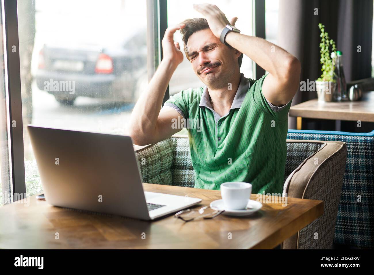 Portrait d'un homme d'affaires désespéré portant un T-shirt vert, se sentant désolé et se blâmant de l'erreur en travaillant en ligne sur un ordinateur portable, prise en intérieur près d'une grande fenêtre, arrière-plan de café. Banque D'Images