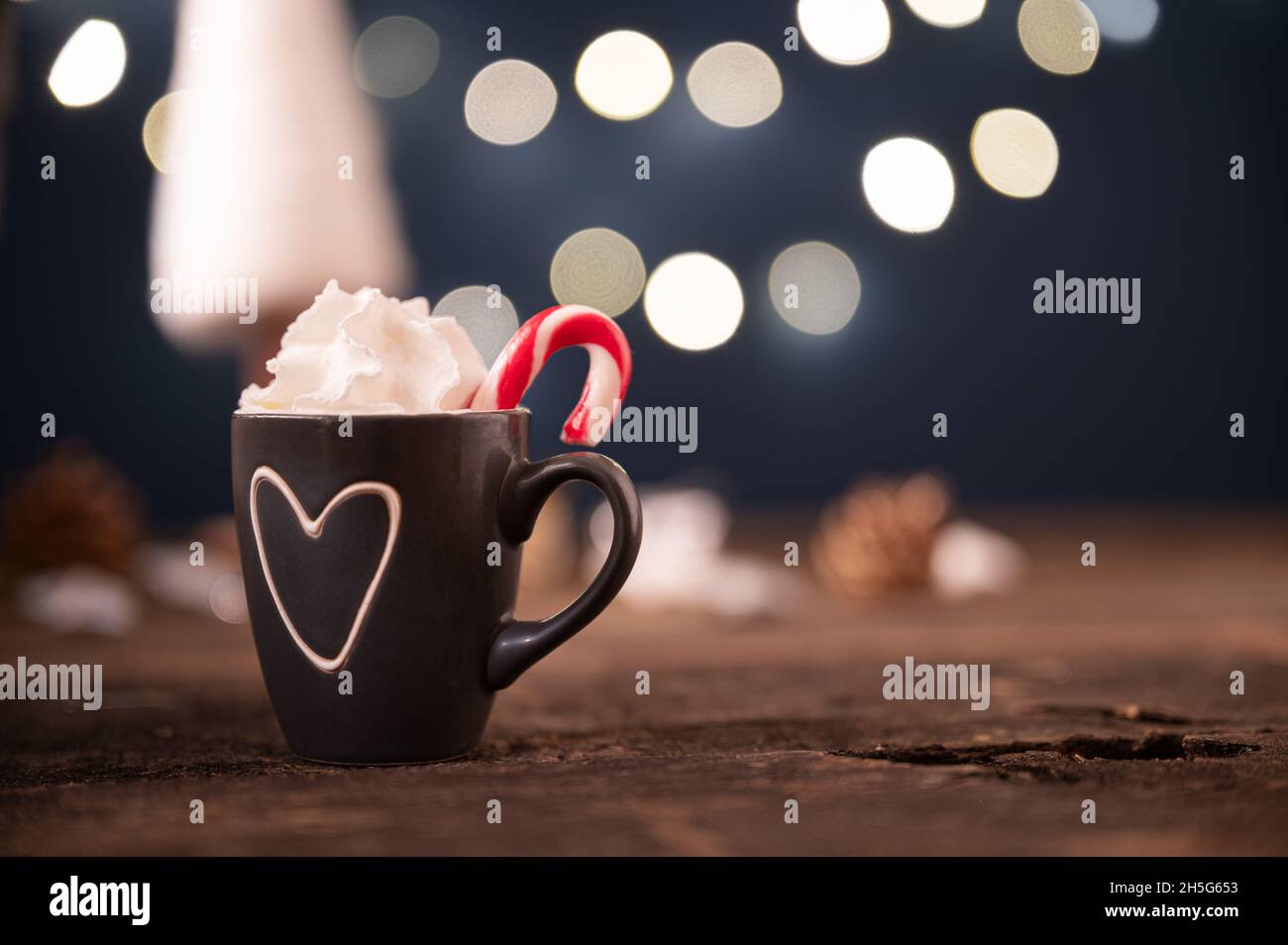 Petite tasse de Noël en forme de coeur pleine de chocolat chaud de vacances avec crème fouettée et lumières de noël floues en arrière-plan. Banque D'Images