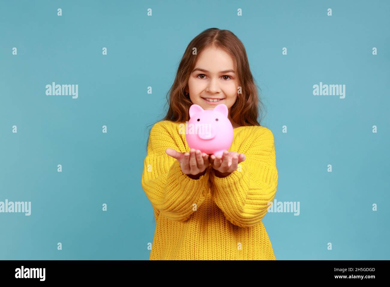 Portrait d'une petite fille souriante tenant une banque de porc à l'appareil photo, heureux d'économiser de l'argent, portant jaune style décontracté chandail.Studio d'intérieur isolé sur fond bleu. Banque D'Images