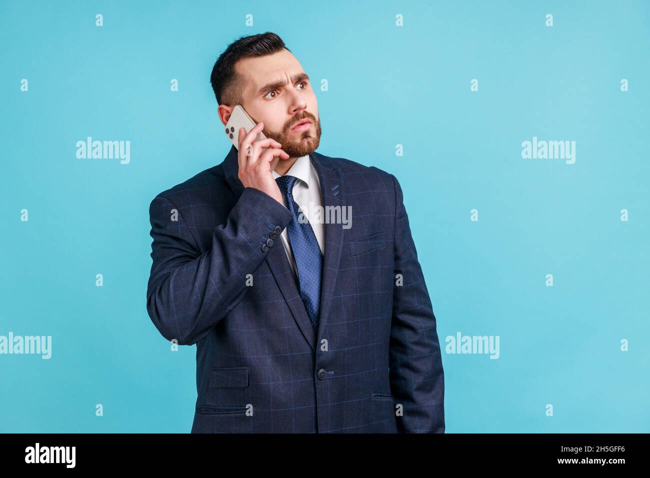 Un homme à barbe sérieux portant un costume de style officiel sombre tenant son téléphone près de l'oreille, parle à ses entreprises, résout des problèmes de travail importants.Studio d'intérieur isolé sur fond bleu. Banque D'Images