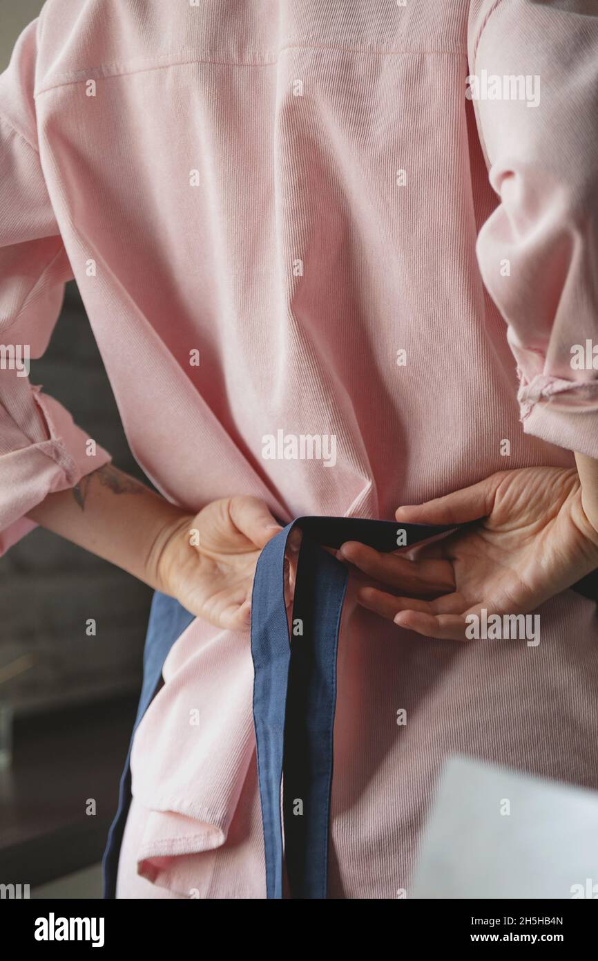Femme en costume rose attache un tablier bleu sur son dos pour commencer à cuisiner dans sa cuisine maison. Image sans visage. Banque D'Images