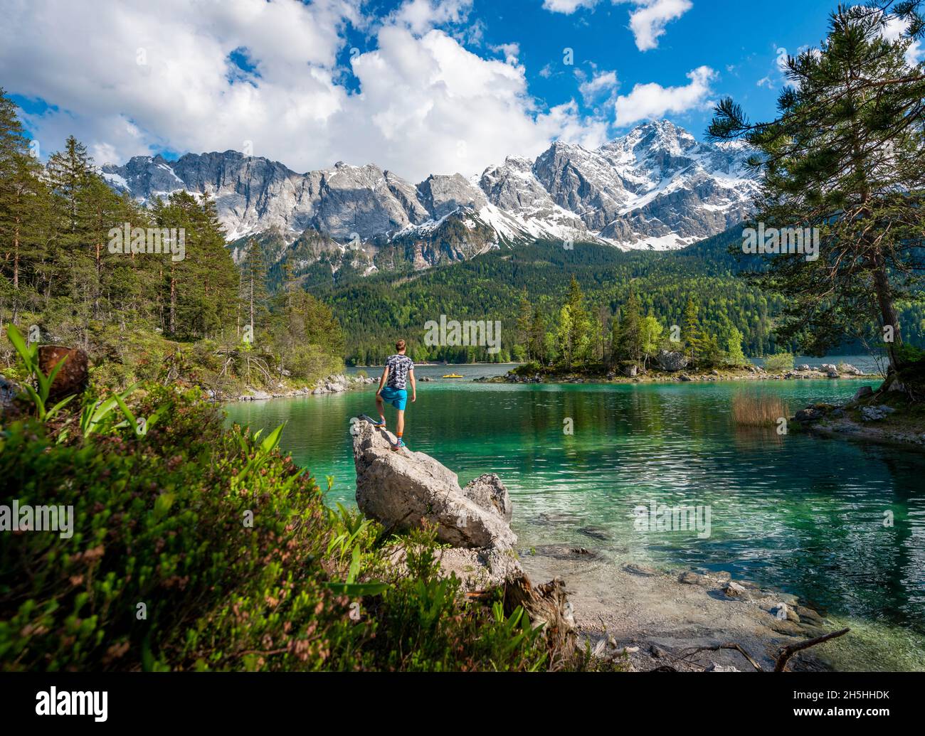 Jeune homme debout sur un rocher, vue sur la distance, lac Eibsee et Zugspitze au printemps avec neige, montagnes Wetterstein, près de Grainau, Upper Banque D'Images