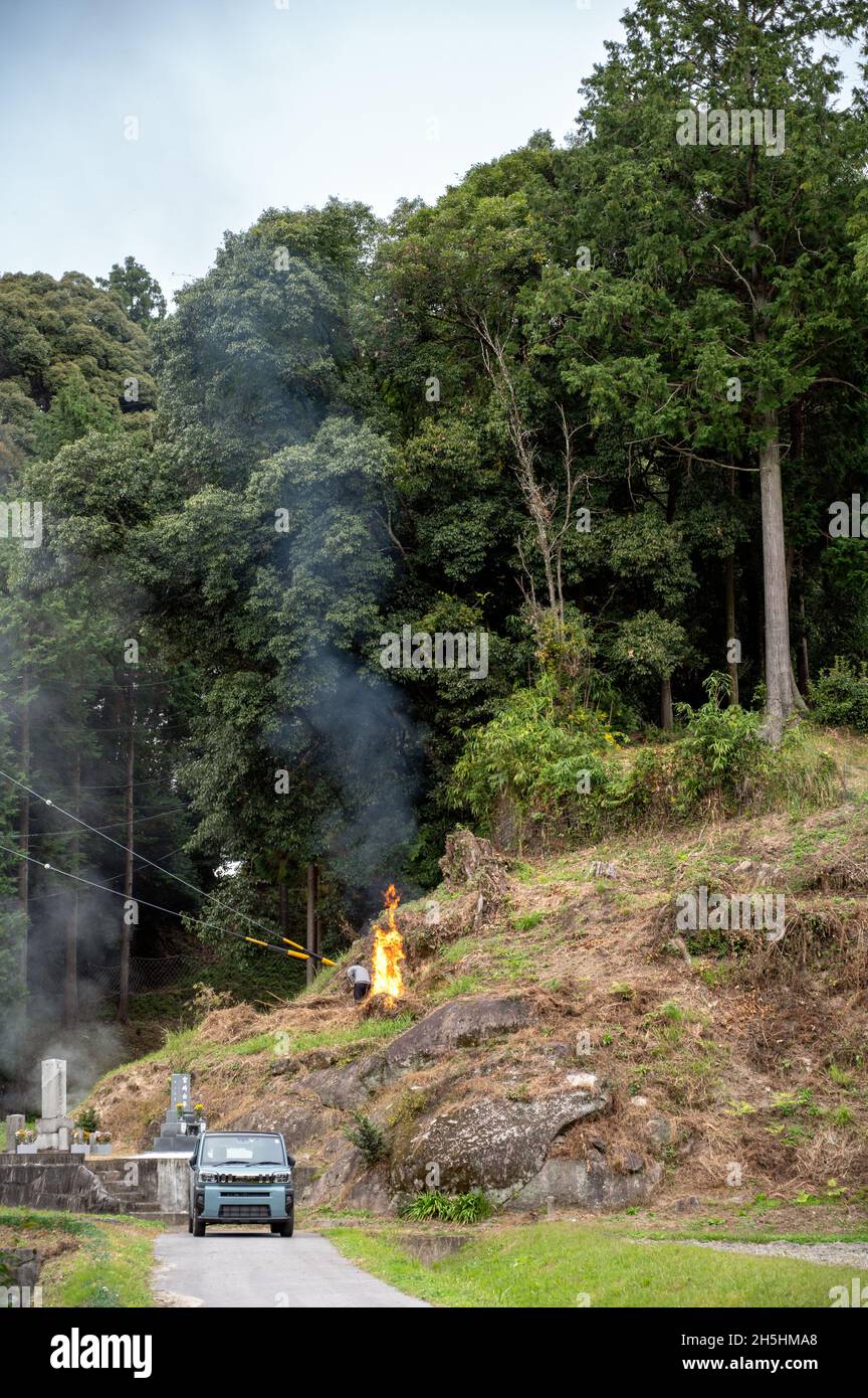 Agriculteur japonais brûlant de l'herbe sèche sur la pente de la montagne.La fumée du feu passe dans l'air.Tombes et une voiture au premier plan. Banque D'Images