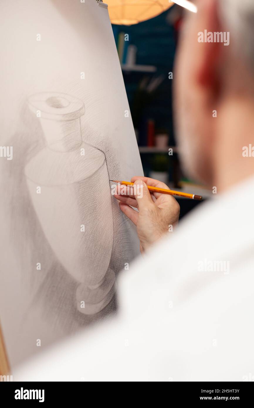 Gros plan de la main caucasienne du vieil homme tenant un crayon sur toile, dessin du modèle de vase au studio d'art.Personne âgée travaillant comme artiste créant un chef-d'œuvre authentique dans l'espace d'atelier Banque D'Images