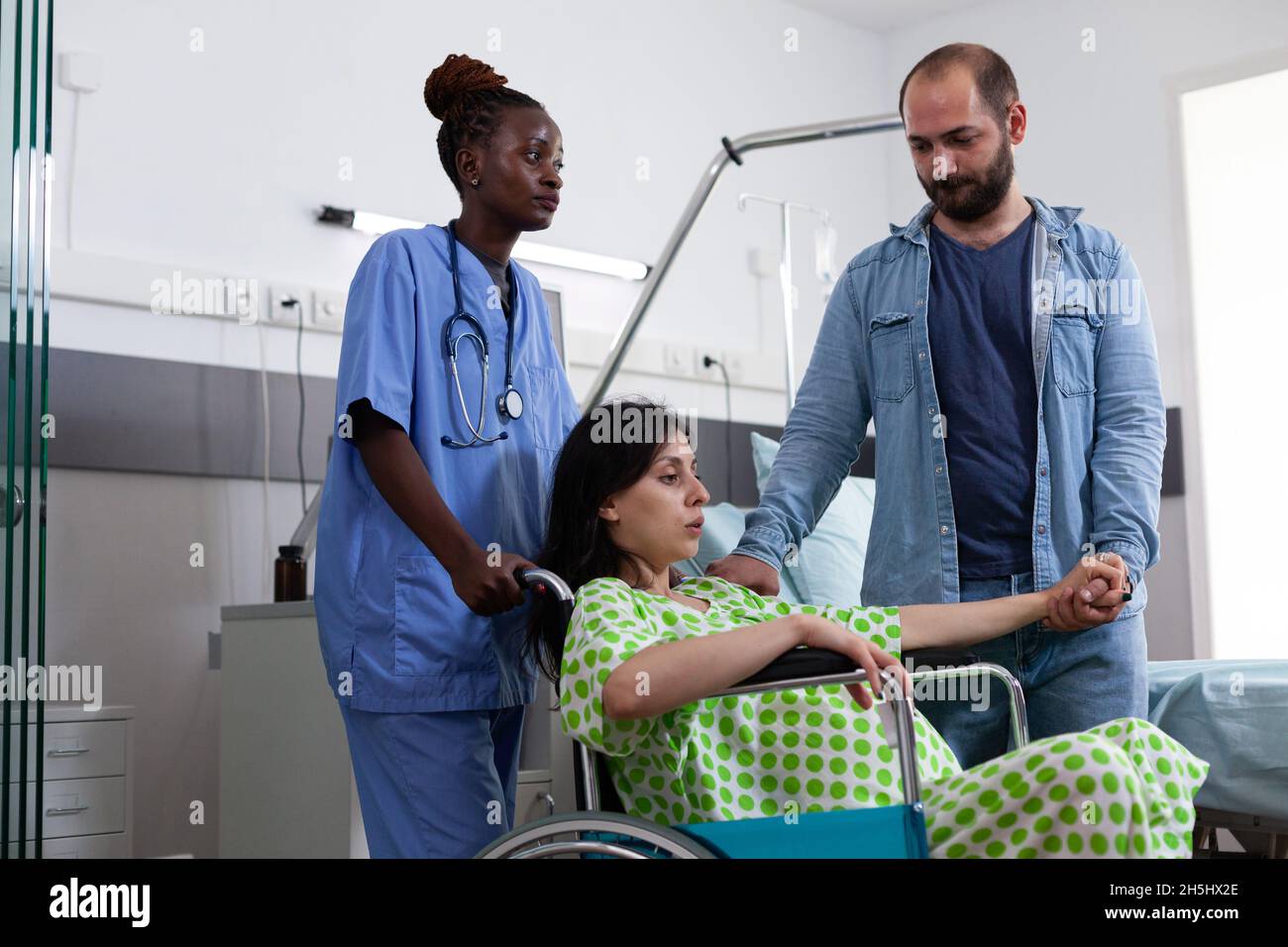 Infirmière noire transportant la femme enceinte en fauteuil roulant vers la salle de maternité pour l'accouchement. Personne avec une bosse de bébé se préparant à l'accouchement tout en tenant la main du mari dans la salle d'hôpital Banque D'Images