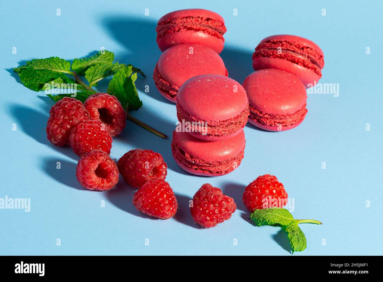 Un assortiment de macarons rouges éclatants à la framboise, accompagnés de framboises fraîches et de feuilles de menthe, disposés sur un fond bleu pastel Banque D'Images