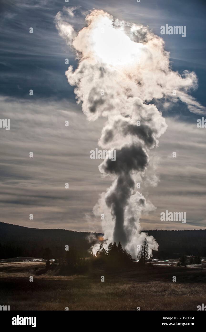 Soleil illuminant une colonne de vapeur thermique en provenance d'Old Faithful en éruption contre un ciel nuageux et bleu et une crête de montagne silhoueted dans le Haut... Banque D'Images
