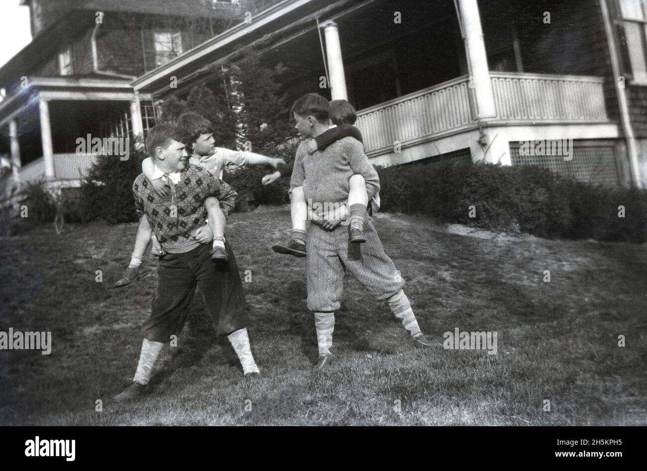 vers 1940, historique, quatre garçons jouant sur une zone herbeuse à l'extérieur de la véranda d'une maison, avec les deux garçons plus âgés portant les deux jeunes garçons sur leur dos.Les garçons plus âgés portent des culottes ou des culottes qui s'étendent juste en dessous du genou et sont portés avec de longues chaussettes.Aussi connu sous le nom de knickerbockeers - ou golfeurs plus ttos, comme ils s'étendent deux pouces sous le genou - ces pantalons de bagardy-kneed étaient un article populaire de vêtements de garçons aux États-Unis au début du XXe siècle. Banque D'Images