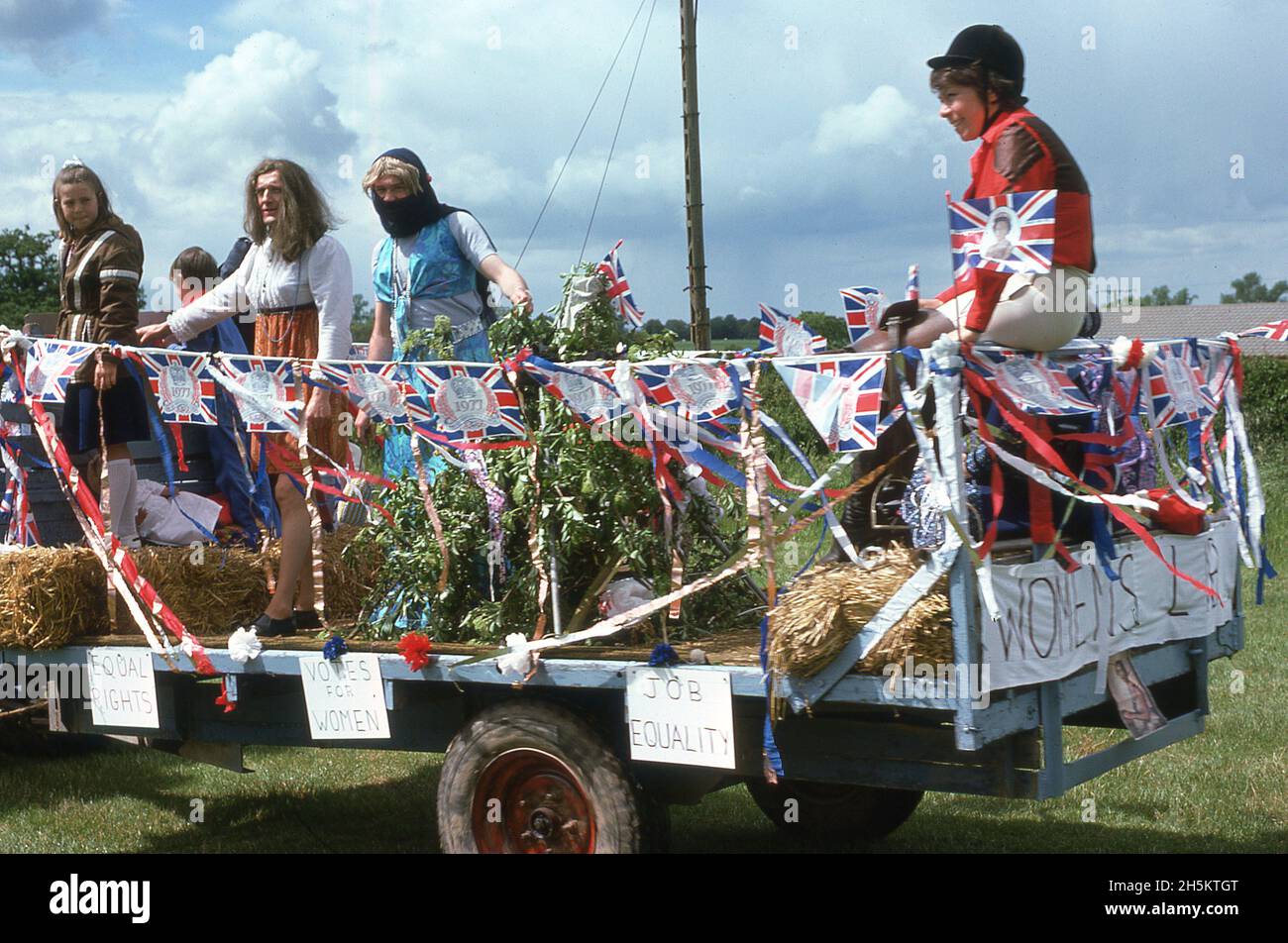 1977, historique, juin et en Grande-Bretagne, les personnes célébrant le jubilé d'argent, du monarque Reine Elizabeth IIÀ l'arrière d'une remorque ou d'un flotteur décoré, deux hommes habillés comme des femmes, plus des filles, un dans une culotte d'équitation assis à l'arrière de la remorque.Les droits des femmes, les votes pour les femmes et l'égalité des droits sont des signes annoncés sur le côté de la bande-annonce, Tilstone, Cheshire, Angleterre, Royaume-Uni. Banque D'Images