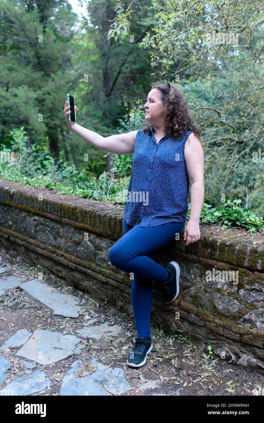Photographie verticale d'une femme caucasienne, avec des cheveux légèrement bouclés, prenant un selfie avec son smartphone dans la forêt.. Banque D'Images