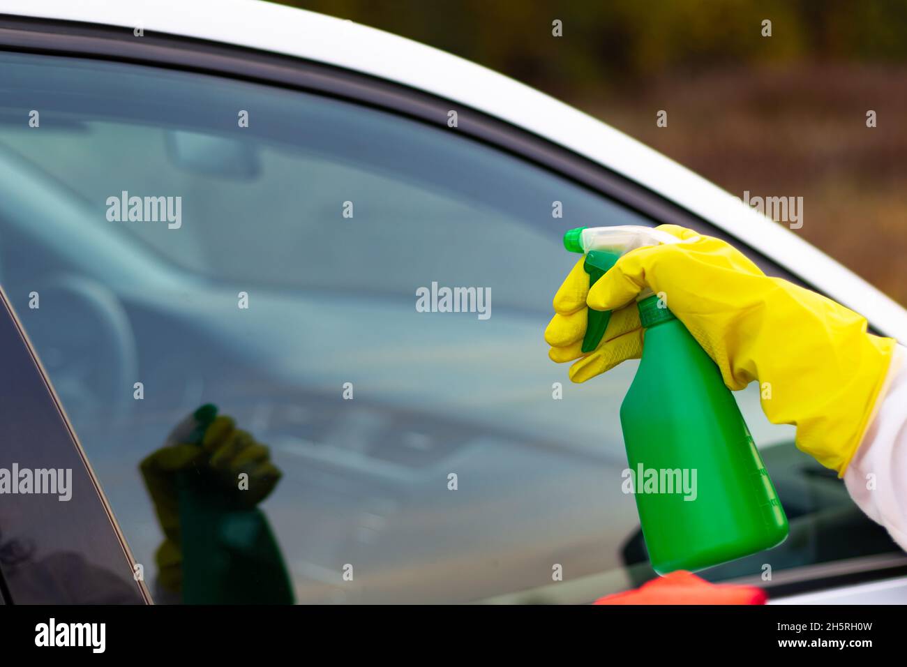 Une main dans un gant en caoutchouc jaune vaporise de l'eau d'une bouteille vaporisée sur une fenêtre de voiture par une chaude journée d'automne.Nettoyage à l'eau.Mise au point sélective.Gros plan Banque D'Images