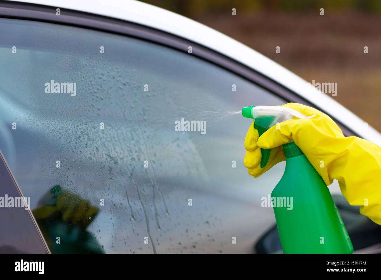 Une main dans un gant en caoutchouc jaune vaporise de l'eau d'une bouteille vaporisée sur une fenêtre de voiture par une chaude journée d'automne.Nettoyage à l'eau.Mise au point sélective.Gros plan Banque D'Images