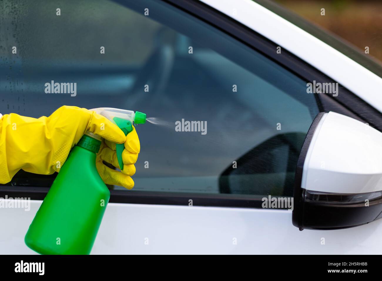 Une main dans un gant en caoutchouc jaune vaporise de l'eau d'une bouteille vaporisée sur une fenêtre de voiture par une chaude journée d'automne.Nettoyage à l'eau.Mise au point sélective.Gros plan Banque D'Images