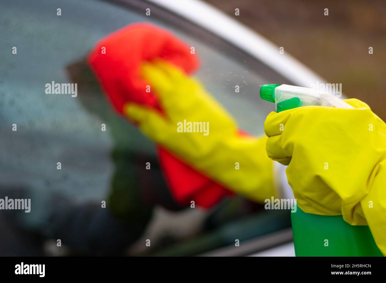 Les mains dans des gants en caoutchouc jaune essuyez le verre d'une voiture avec un chiffon rouge et vaporisez de l'eau à partir d'un pistolet à pulvérisation par une chaude journée d'automne.Nettoyage à l'eau.Sélectif FO Banque D'Images