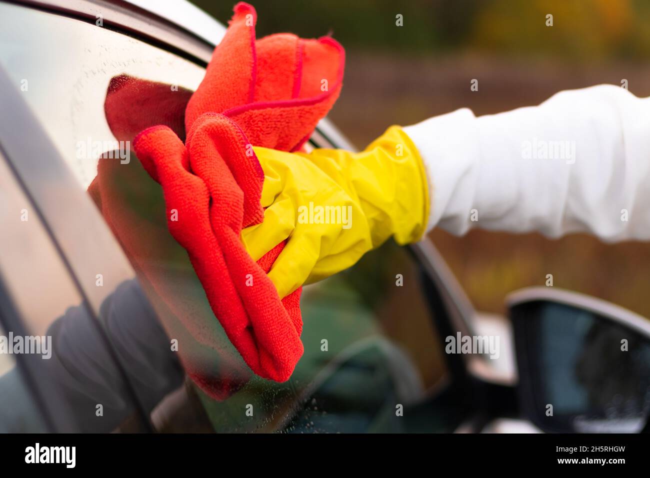 Une main dans un gant en caoutchouc jaune essuie le verre d'une voiture avec un chiffon en microfibre rouge de la saleté lors d'une chaude journée d'automne.Nettoyage à l'eau.Mise au point sélective.Clos Banque D'Images