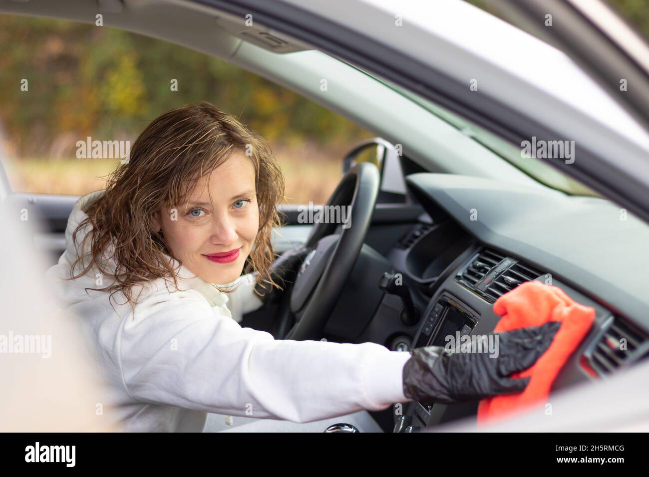 Une belle jeune femme pilote dans une veste blanche et des gants noirs essuie la poussière à l'intérieur de la voiture avec un chiffon rouge lors d'une chaude journée d'automne.Sélection Banque D'Images