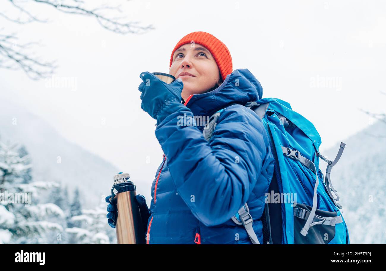 Femme d'âge moyen souriant et buvant une boisson chaude de thermos ballon habillé chaud veste en bas pendant qu'elle trekking route des montagnes d'hiver. Peopl. Active Banque D'Images