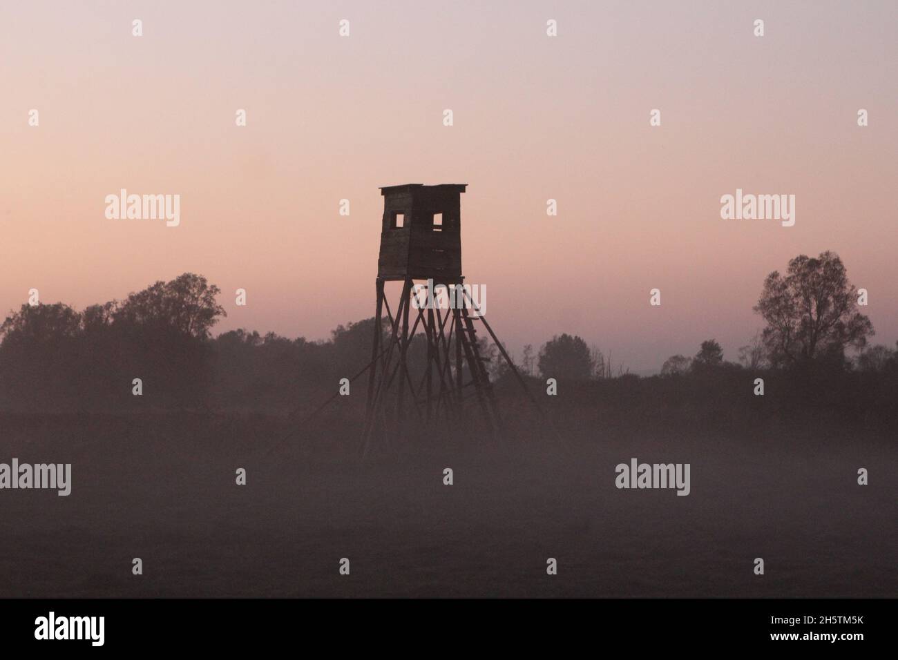 Überdachte Ansitzeinrichtung als Hochsitz (Ansitz) aus Holz zur Jagdausübung auf einem Feld im Nebel in der rötlichen Abenddämmerung.Rügen. Banque D'Images