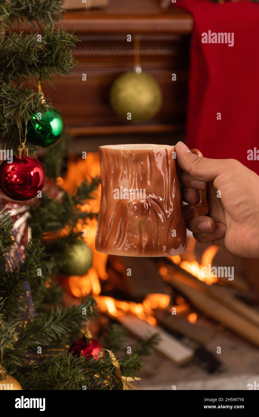 main tenant une tasse originale avec un design en bois, avec une boisson chaude, en arrière-plan une cheminée en bois avec une flamme, appréciant à la maison en hiver Banque D'Images