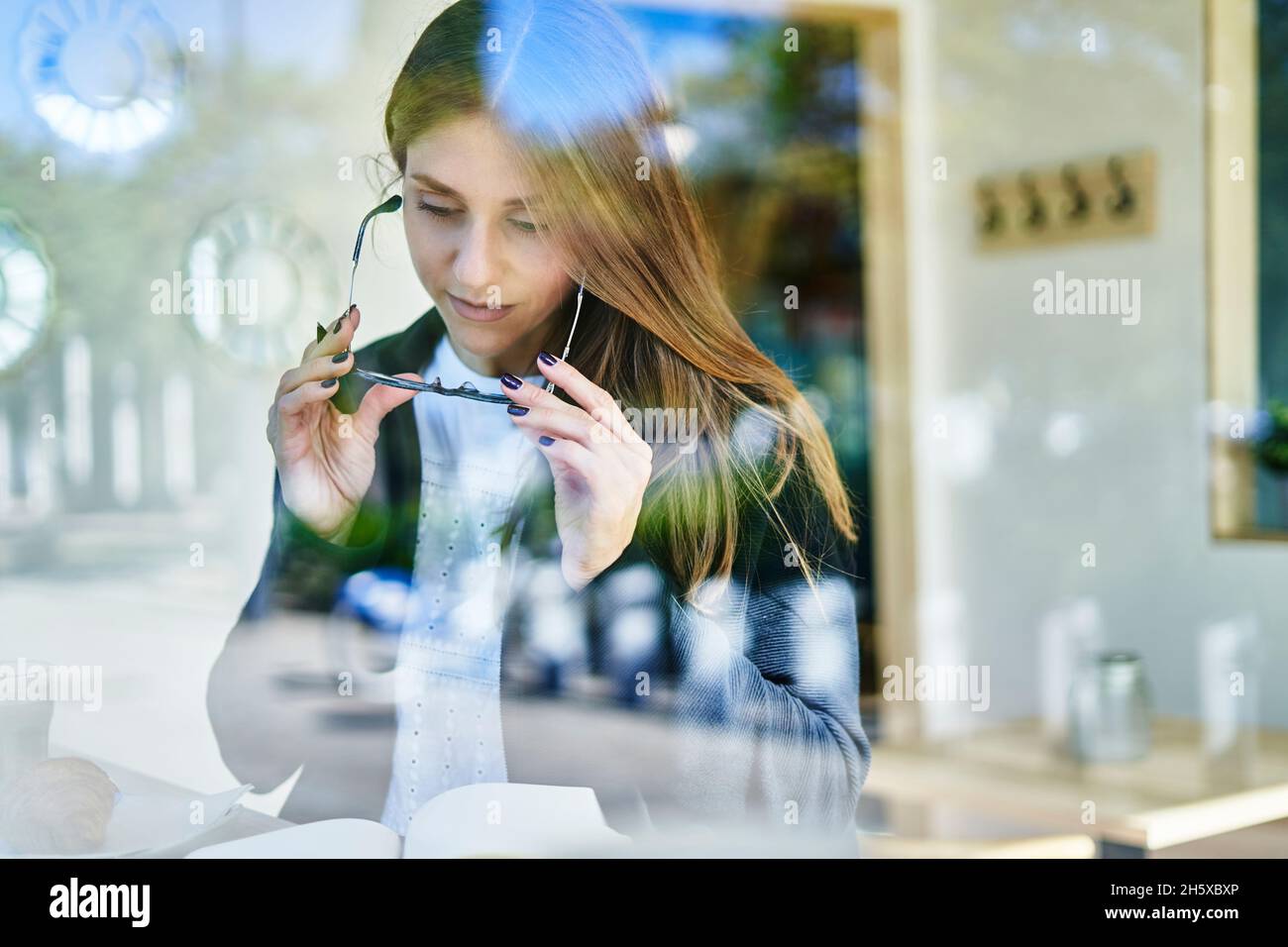 À travers le verre de la travailleuse concentrée prenant des lunettes tout en lisant des notes dans le bloc-notes à la table avec café et croissant dans le café Banque D'Images