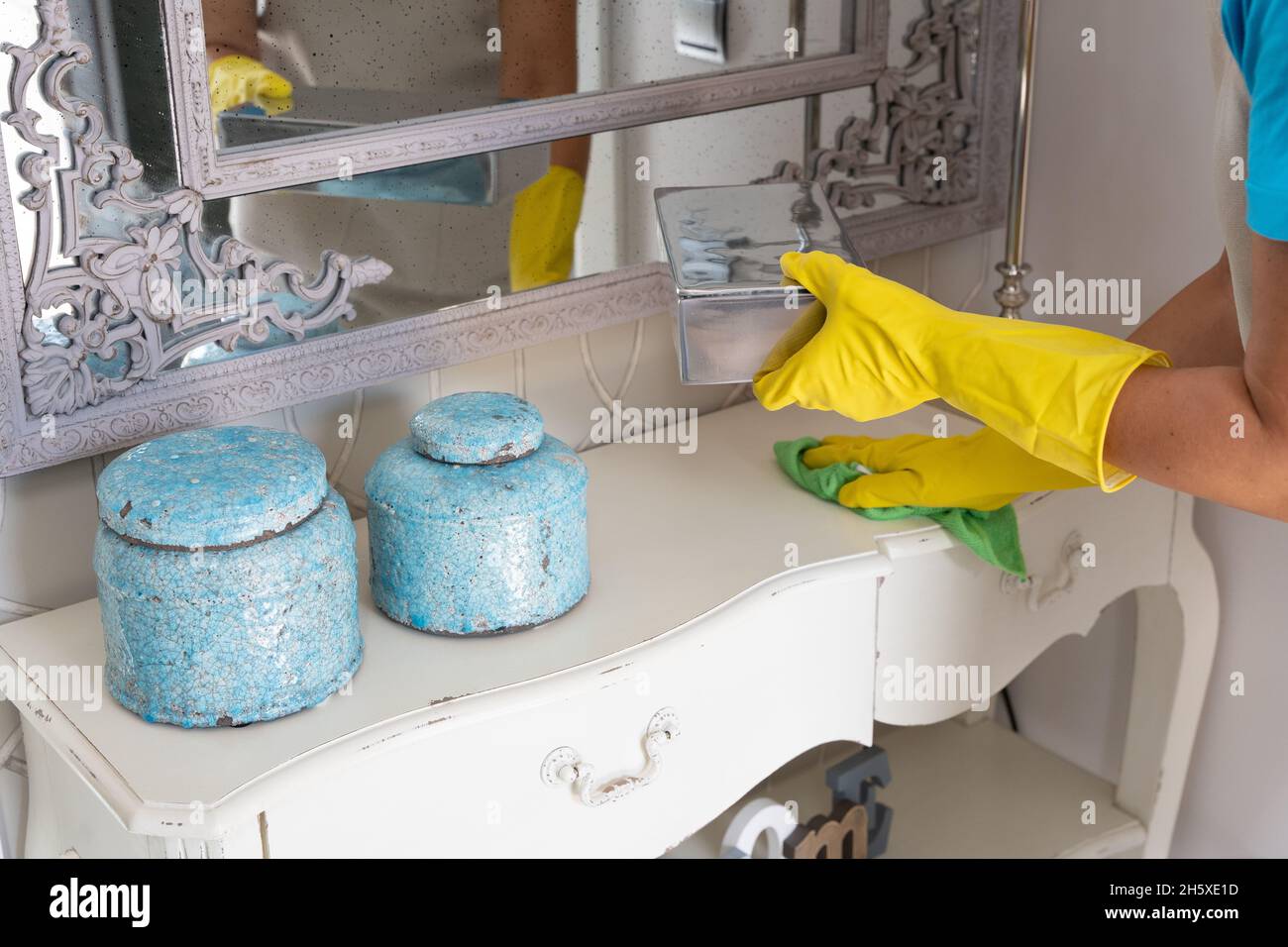 Récolte inreconnaissables femme femme gouvernante dans un tablier et des gants en caoutchouc essuyant les éléments décoratifs placés sur l'armoire sous le miroir dans le hall pendant le travail dans eleganan Banque D'Images