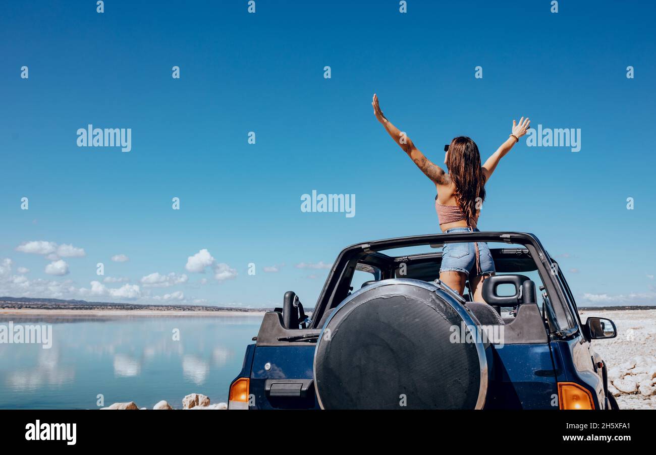 Vue arrière d'une jeune femme en haut et d'un short en denim, les bras relevés dans une voiture de sport décapotable garée sur un sable fin Banque D'Images