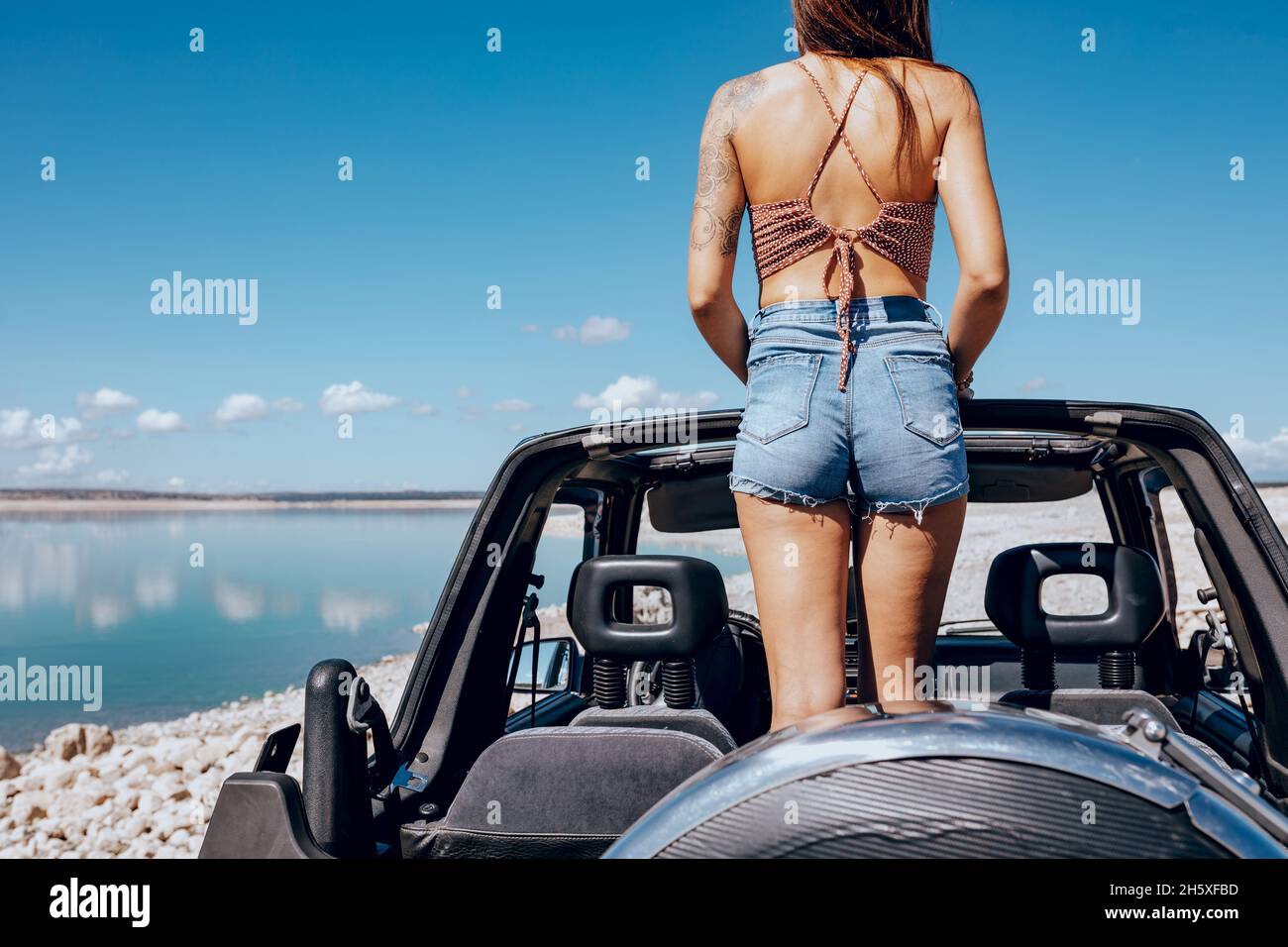 Vue arrière d'une jeune femme en haut tatouée et d'un short en Jean qui se trouve dans une voiture de sport convertible garée sur une plage de sable et admirant Banque D'Images