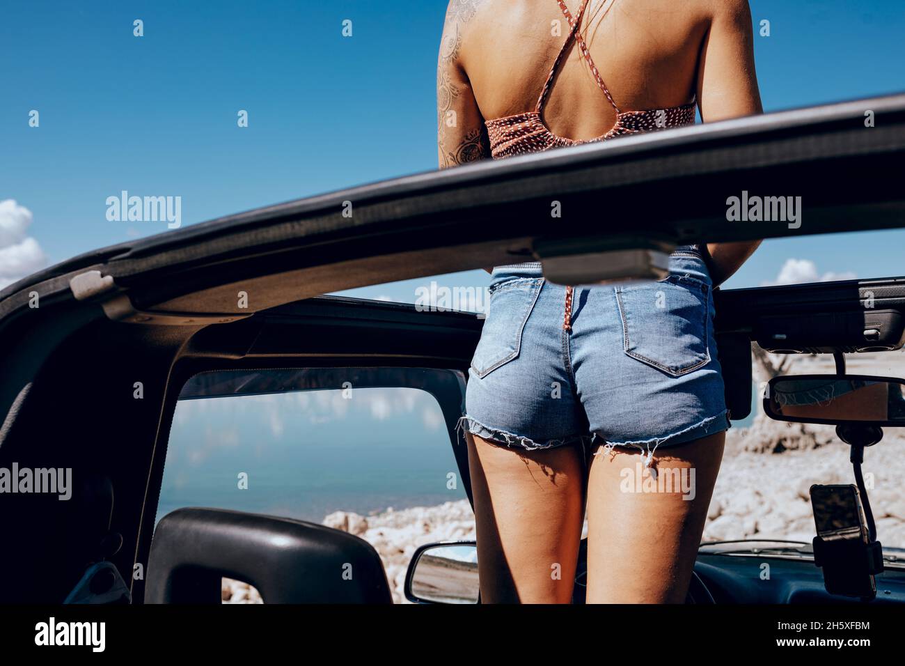 Vue arrière d'une jeune femme en haut tatouée et d'un short en Jean qui se trouve dans une voiture de sport convertible garée sur une plage de sable et admirant Banque D'Images