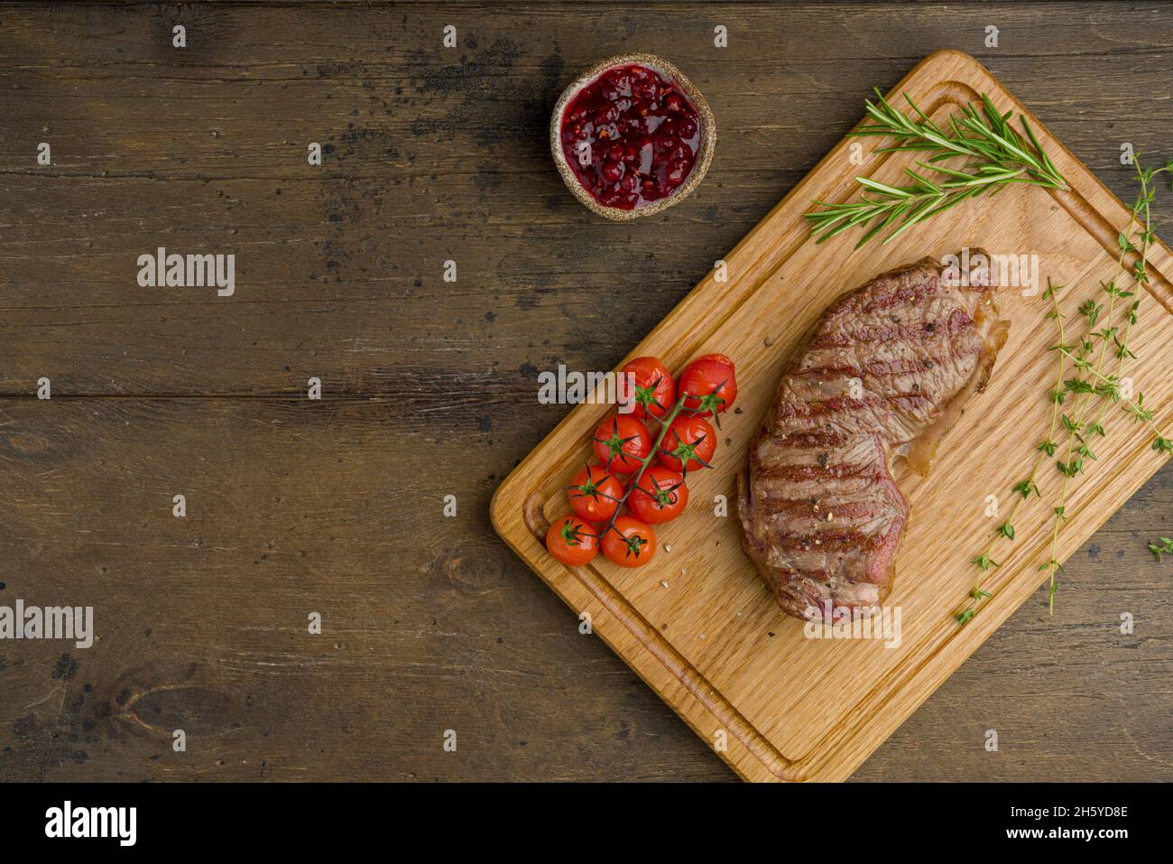 Steak de stripine grillé avec tomates cerises, thym, romarin et sauce aux baies de lingonis sur planche à découper en bois.Délicieuse viande juteuse et tendre.Horizont Banque D'Images