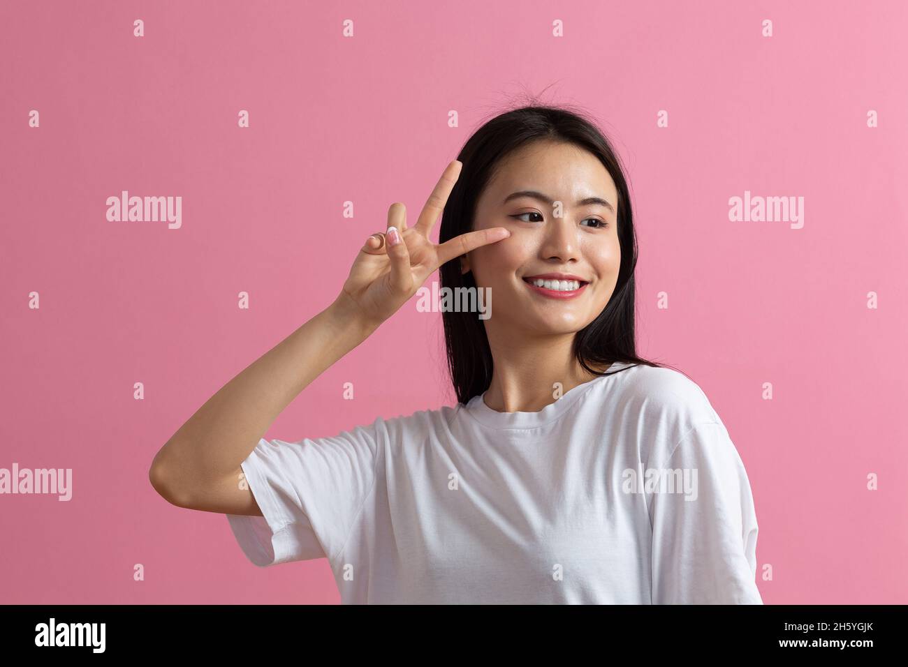 Portrait de la jeune femme souriante positive asiatique avec geste des doigts en V sur fond rose. Banque D'Images