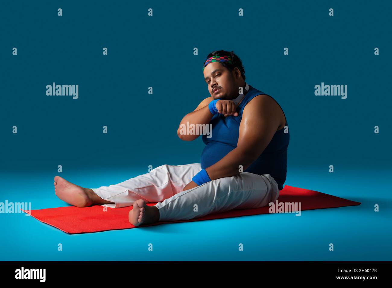 Un gros homme assis sur un tapis de yoga essuyant sa sueur épuisée après un entraînement. Banque D'Images