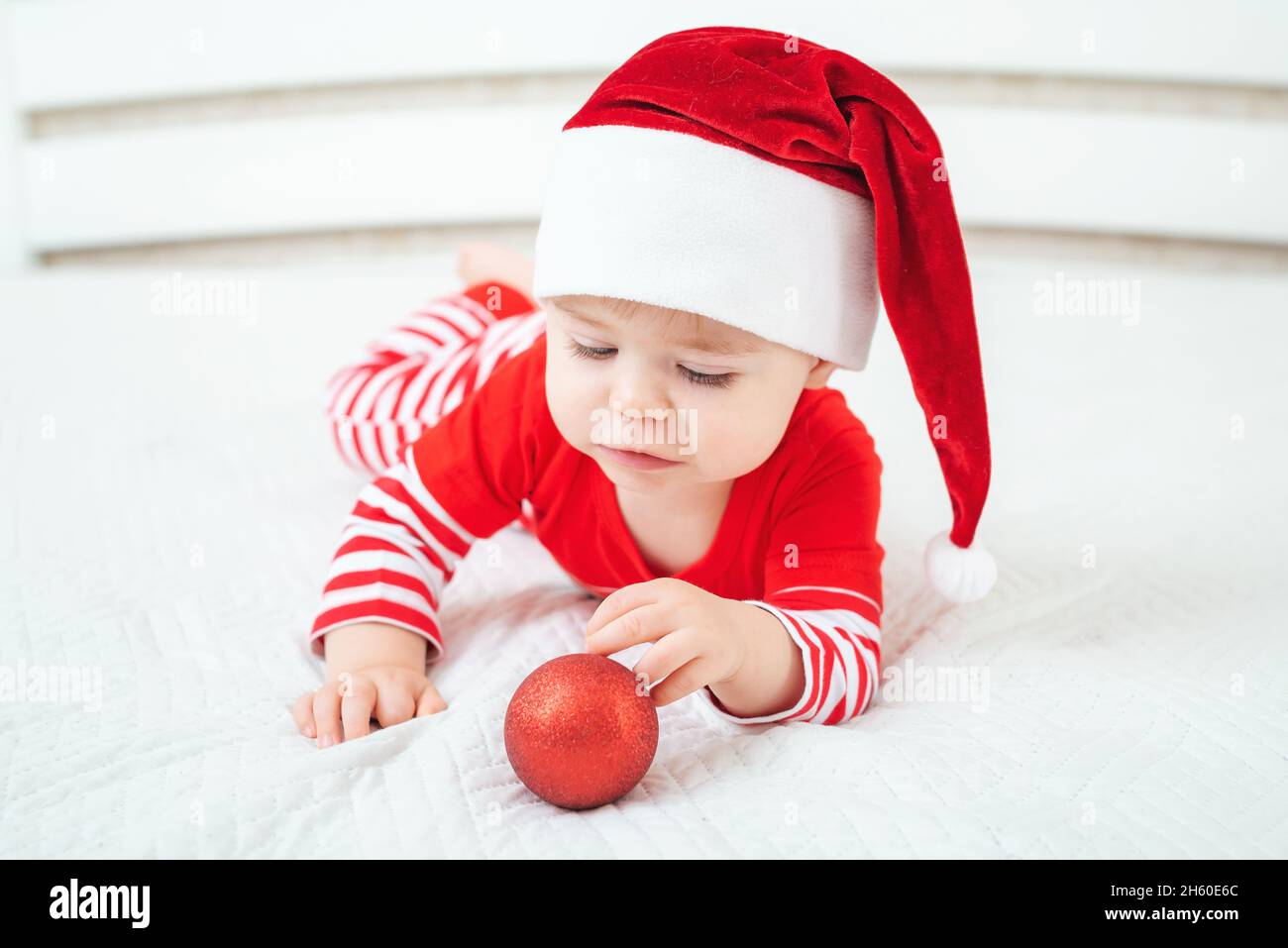Un bébé d'un an en chapeau de père Noël repose sur le ventre et joue avec  le ballon de Noël écarlate.Enfant portant un costume rayé rouge blanc.Joyeux  Noël et heureux Photo Stock -
