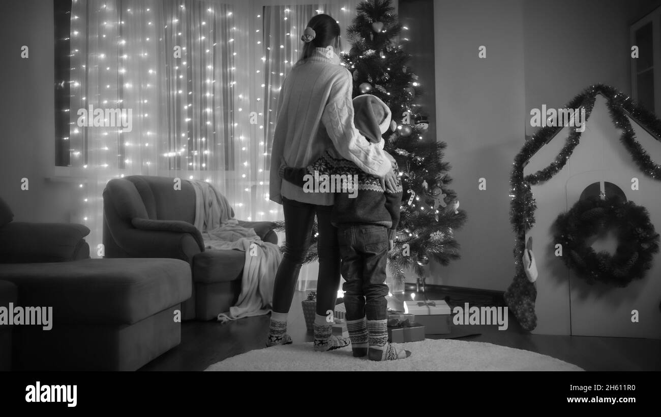 Photo en noir et blanc de maman avec son fils dans un chapeau de père Noël debout à côté de l'arbre de Noël lumineux dans la salle de séjour Banque D'Images