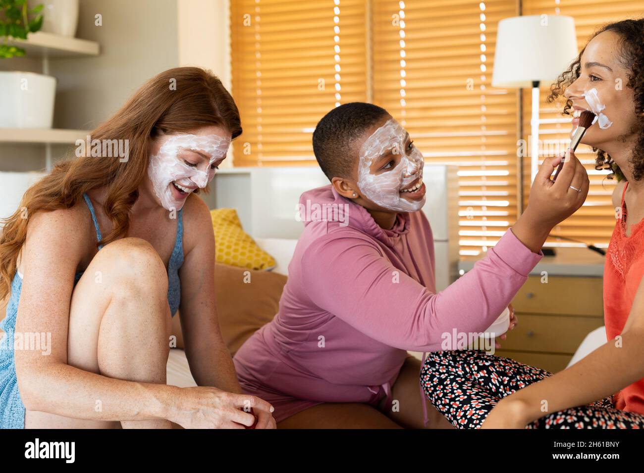 Des amies biraciales souriantes avec masques faciaux assises à la maison pendant le week-end Banque D'Images