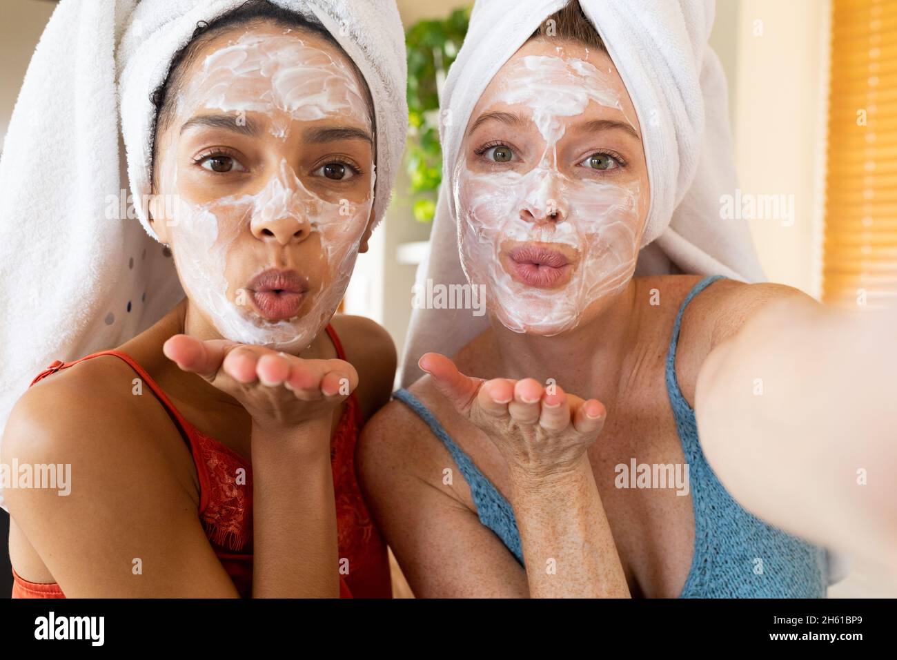 Portrait d'amis femelles avec masques faciaux et serviettes enveloppées sur des baisers soufflants de cheveux à la maison Banque D'Images