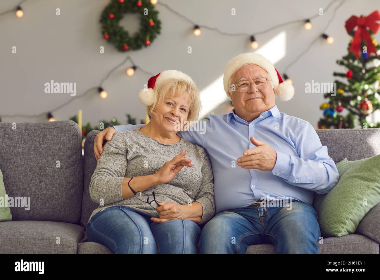 Couple senior souriant en chapeaux de Noël assis sur le canapé et regardant l'appareil photo pendant la vidéo en ligne Banque D'Images