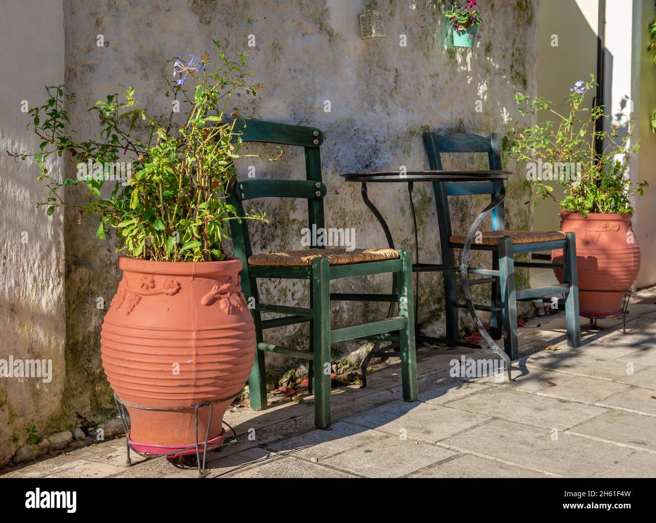 Une table et des chaises près du mur extérieur d'une maison de village grecque avec des plantes dans de grands pots des deux côtés. Banque D'Images