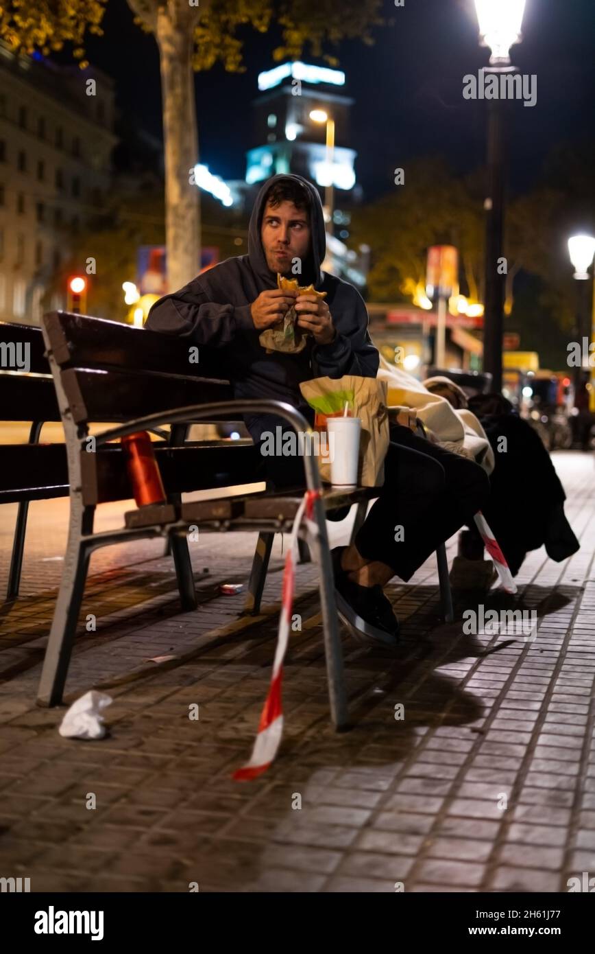 Un homme sans-abri dans un hamburger à capuchon et en regardant loin tout en étant assis sur le banc dans une rue éclairée la nuit Banque D'Images