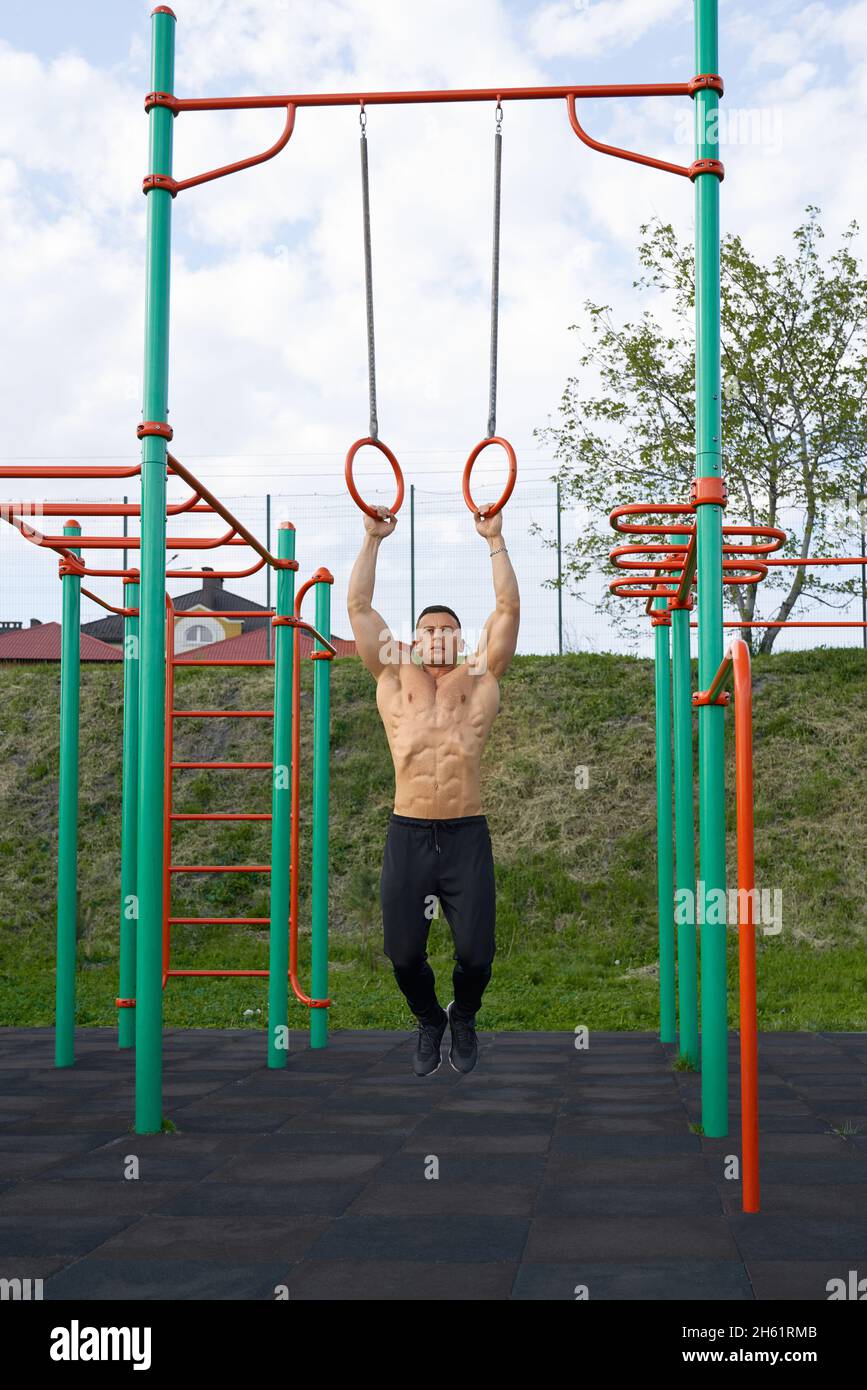 Homme sans chirless fort faisant des pull-ups sur les anneaux de gymnastique à l'extérieur.Caucasien bodybuilder avec corps musculaire enjoung entraînement sur terrain de sport. Banque D'Images