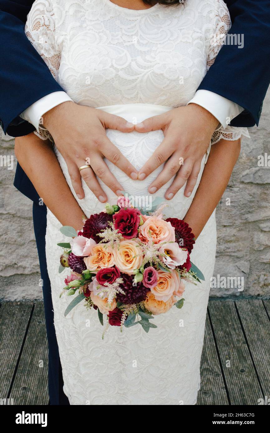 femme enceinte en robe de dentelle blanche avec le bouquet de mariée coloré, le mari tient le ventre de grossesse Banque D'Images