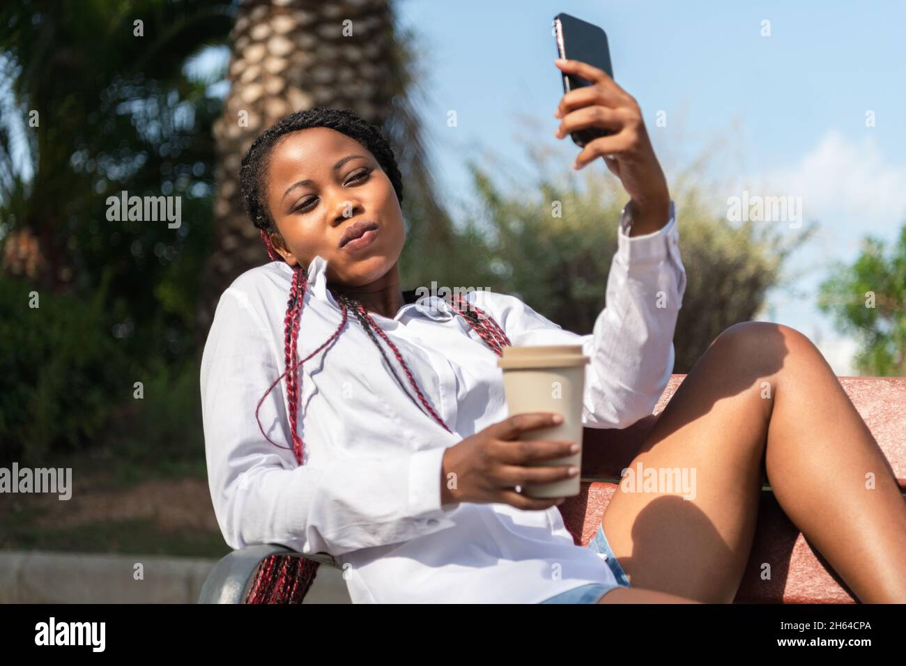 Jeune femme assise sur un banc prenant un selfie et tenant un café à emporter Banque D'Images