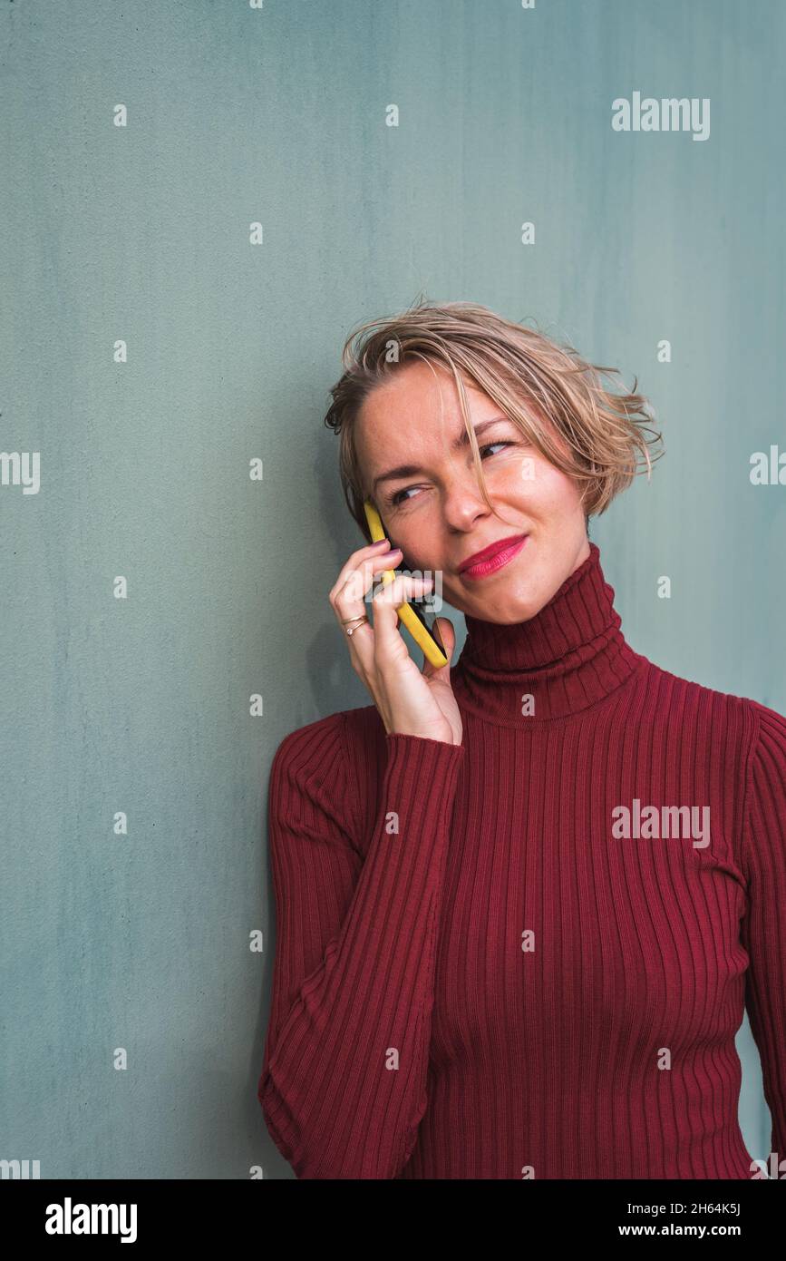 portrait vertical d'une femme blonde sur un chandail en grenat parlant au téléphone tout en s'appuyant sur un mur vert Banque D'Images