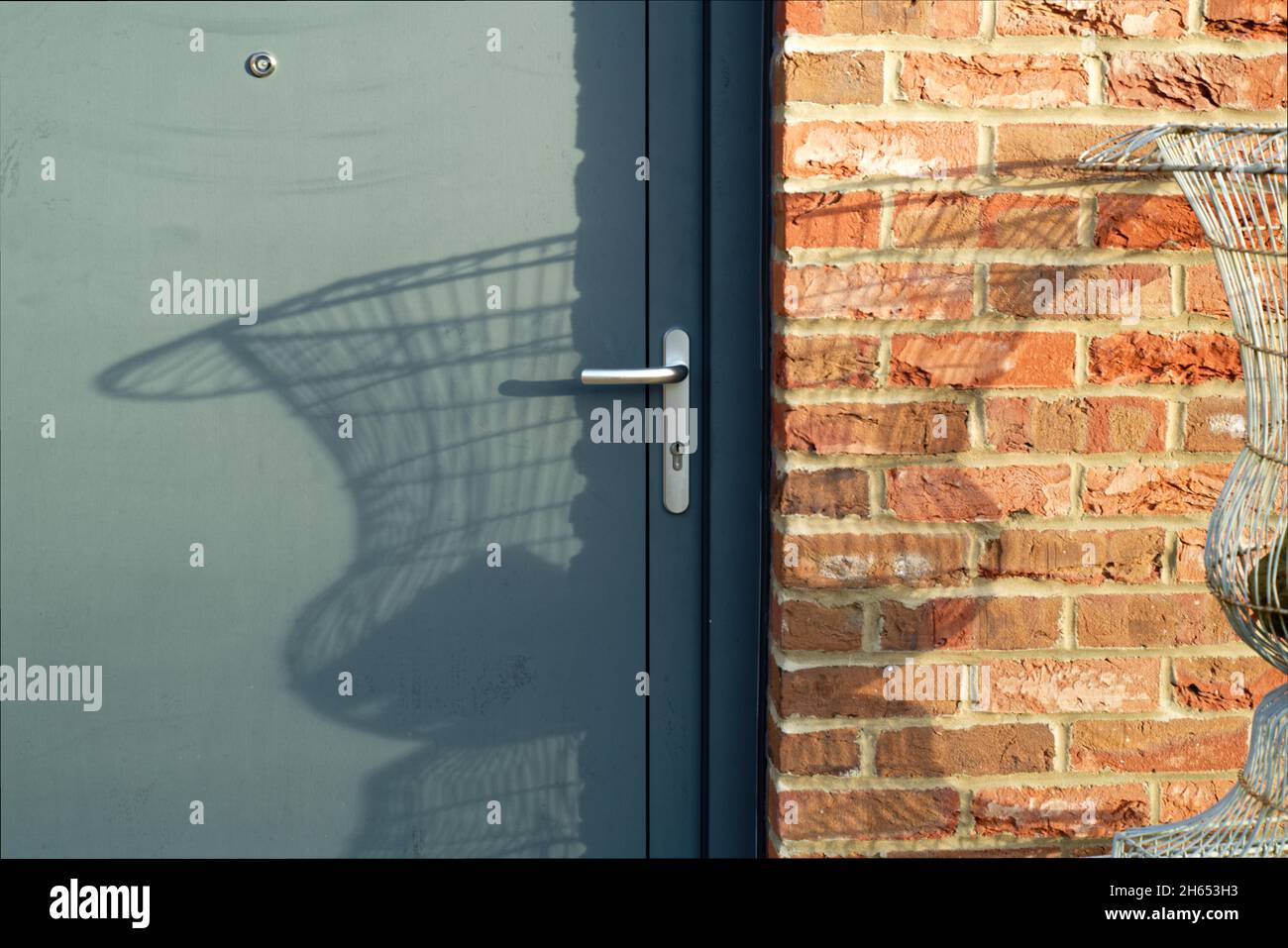 L'ombre faite par un panier en fil de fer/semoir urent contre un mur en brique et la porte par la lumière du soleil forte du matin. Banque D'Images