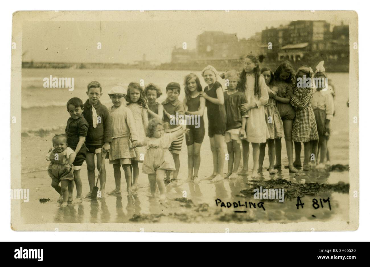 Original charmante époque édouardienne début des années 1900 carte postale d'enfants petits et plus âgés pagayant au bord de la mer, formant une ligne pour le photographe - plus petit enfant à l'avant. Ils portent une sélection de vêtements et de costumes de bain, Margate jetée / jetée est en arrière-plan. Photographié sur la plage en contrebas de Margate Parade, Margate, Kent. Circa 1910 rétro photo de plage. Banque D'Images