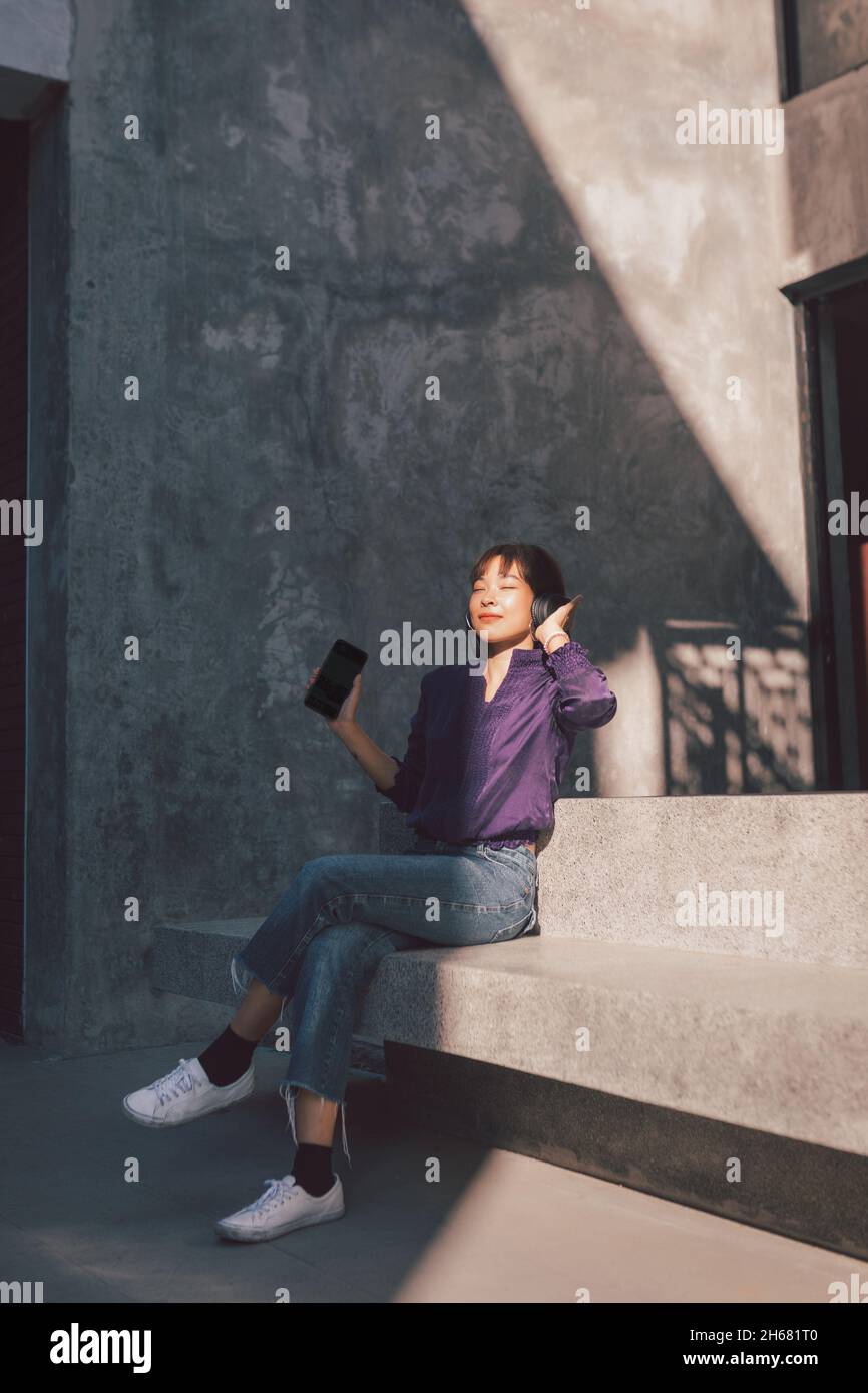 Une jeune femme asiatique heureuse écoute de la musique avec un casque via un smartphone et s'amuse en étant assise dans la rue. Banque D'Images Une jeune femme asiatique heureuse écoute de la musique avec un casque via un smartphone et s'amuse en étant assise dans la rue. Banque D'Images