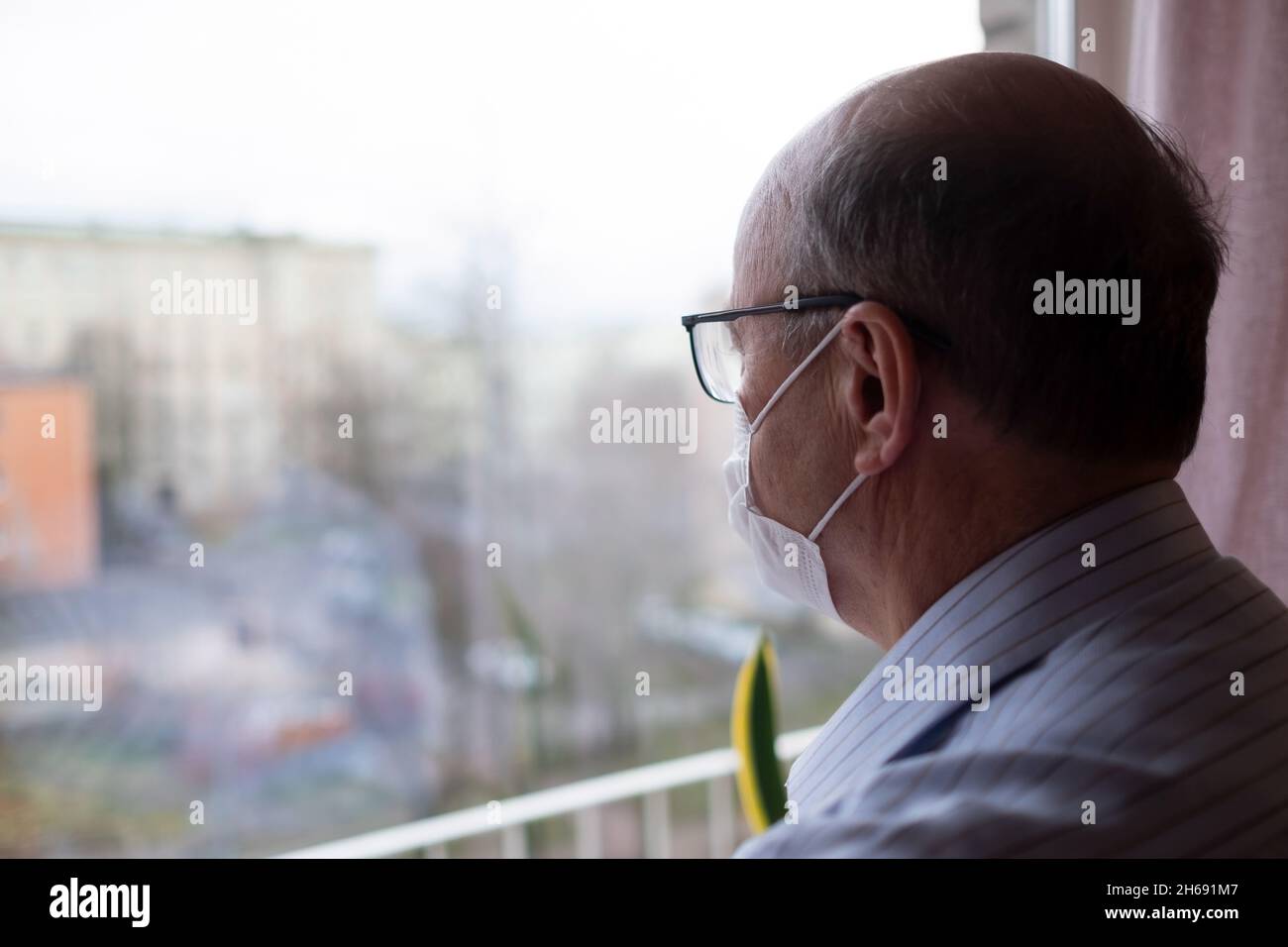 Homme âgé de race blanche portant un masque facial regardant dans la rue près de la fenêtre Banque D'Images