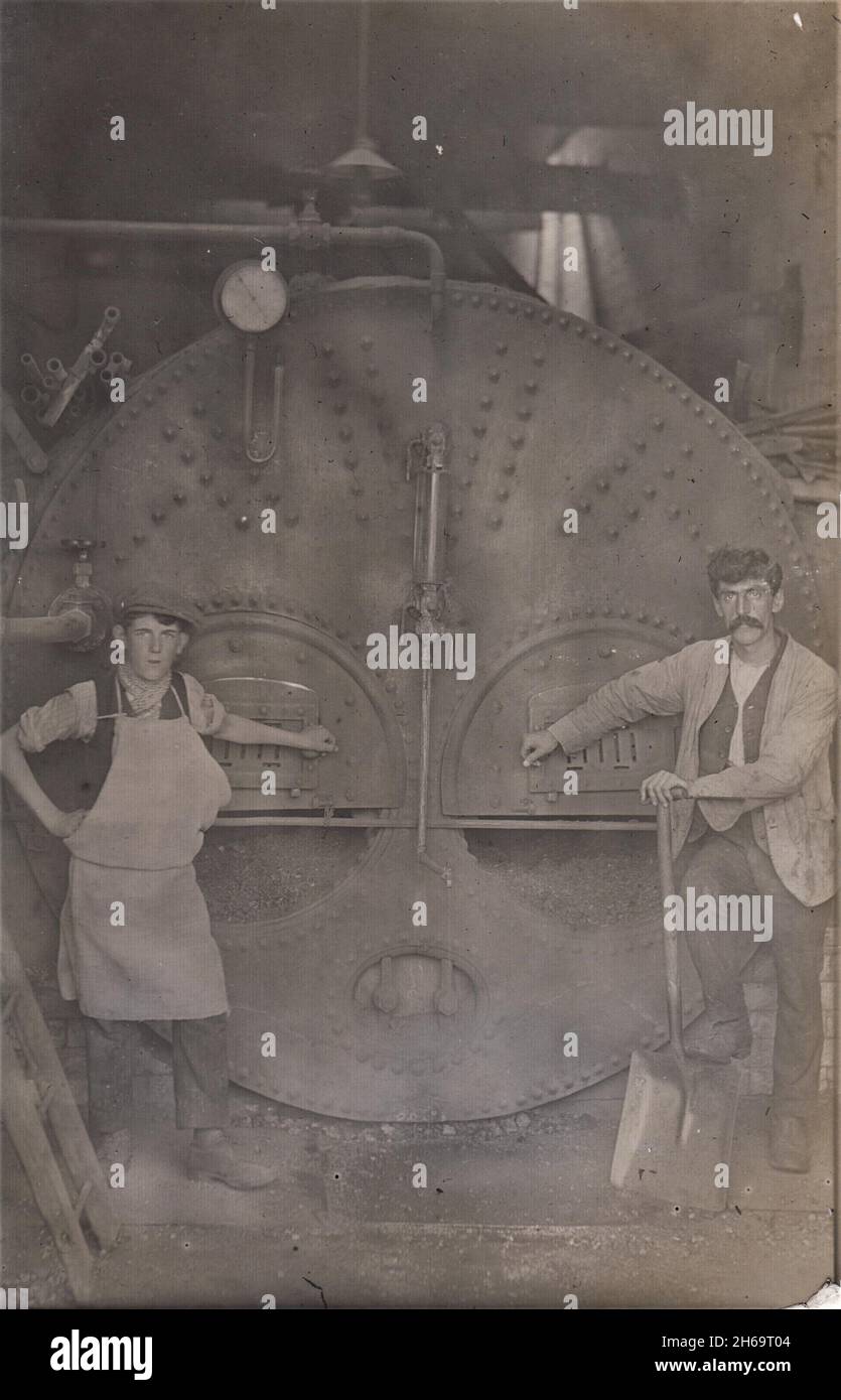 Deux moteurs ou cuiseurs debout à côté du four de la chaudière d'un moteur à vapeur stationnaire dans une usine ou une usine. L'homme plus âgé est debout avec une bêche, le jeune homme porte un tablier, tous deux ont leurs mains sur les portes de la machine industrielle, début 20th siècle Banque D'Images