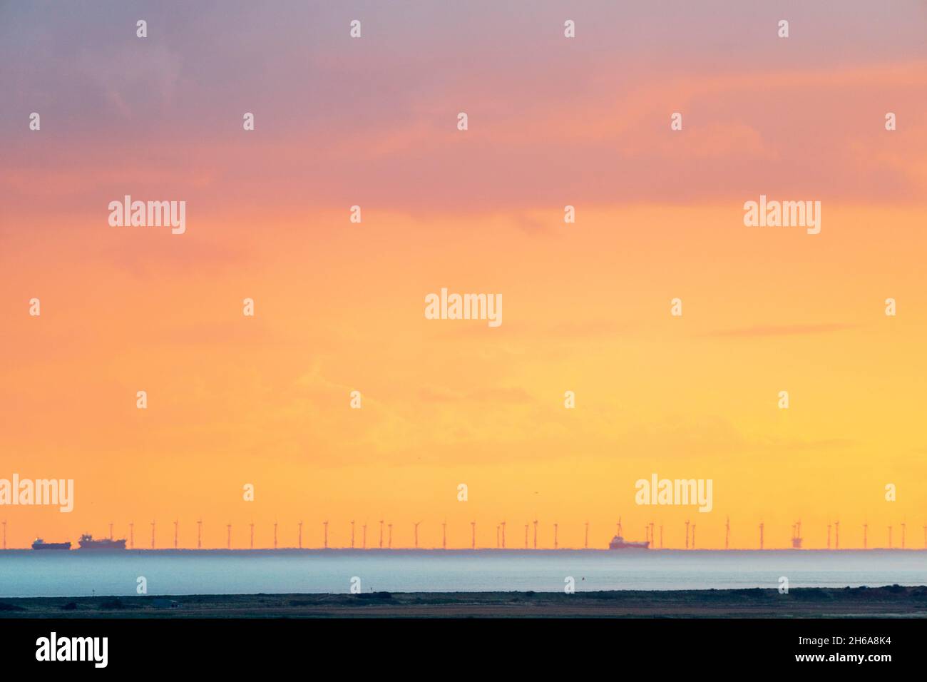 Manche avec un ciel de lever de soleil orange nuageux, un horizon bas, des navires de cargaison et des éoliennes d'un parc éolien offshore vu par une forte brume de chaleur. Banque D'Images