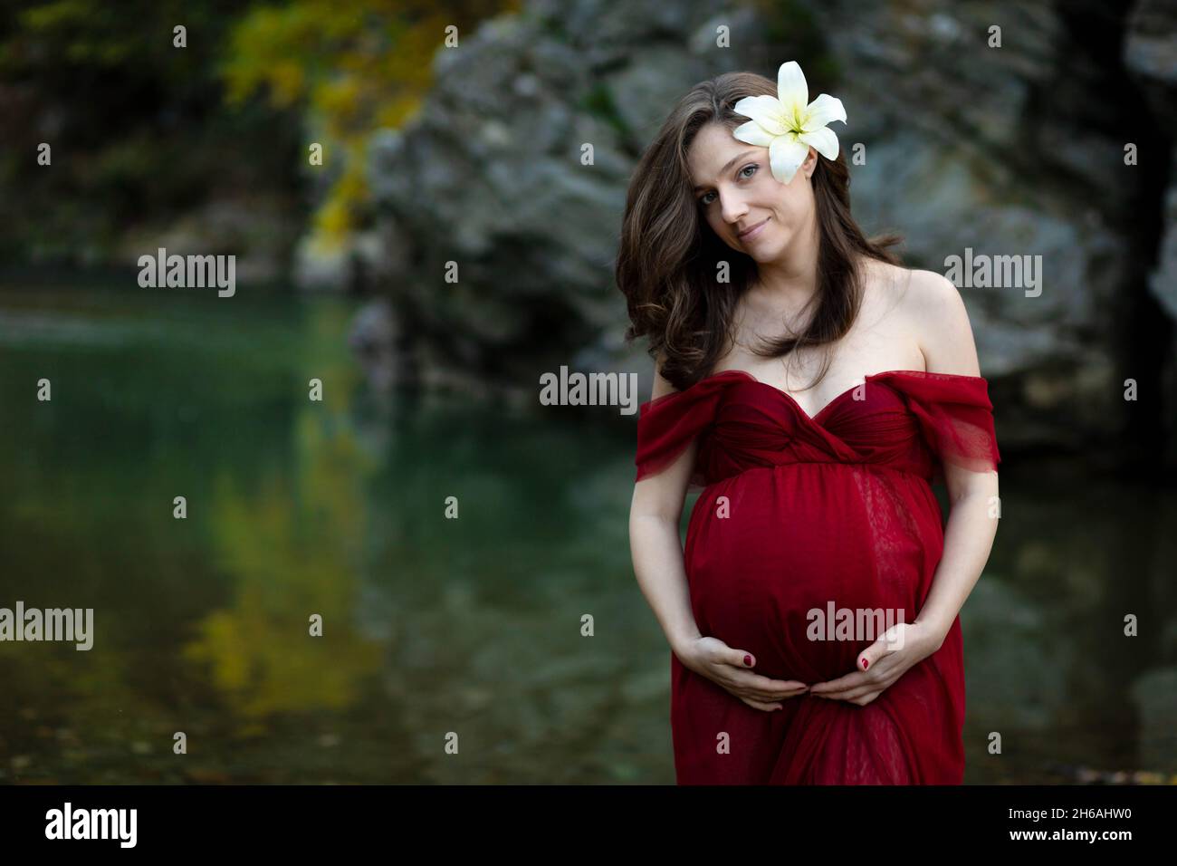 Jeune femme enceinte blonde, élégante, debout dans la rivière en robe rouge, fleur blanche derrière son oreille Banque D'Images